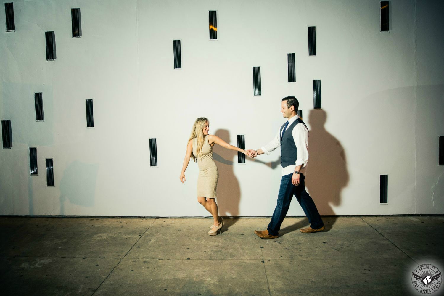 Hot, fit, petite  blond woman in a tight tan ribbed dress and tan heels holds hand with a brunette guy wearing a white untucked button up shirt with a black skinny tie, a dark grey vest, dark blue jeans and brown dress shoes while walking happily in front of white wall with colored glass blocks in this monochromatic  engagement photo at the Contemporary  Austin.